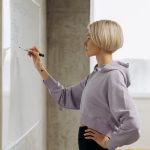 Fit female trainer writes workout plan on a whiteboard in a gym setting. Side view.